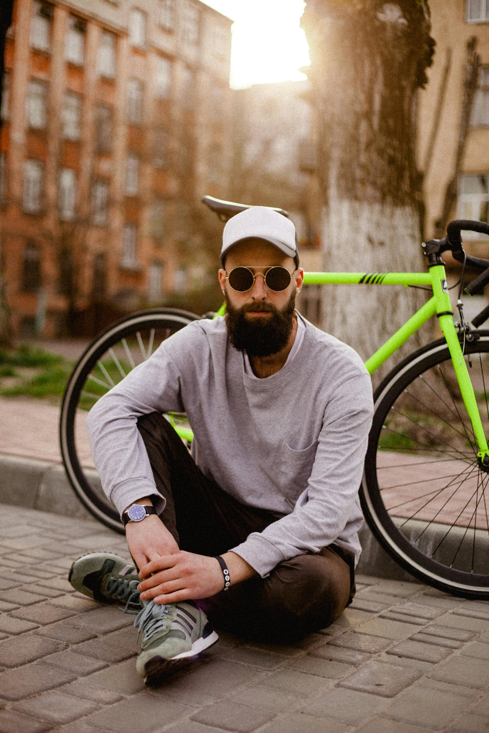 Fashionable man with sunglasses and beard sitting next to a bicycle on an urban street.